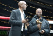 Two men stand side by side, each holding a trophy, smiling proudly at the camera.