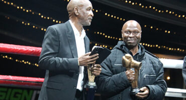 Two men stand side by side, each holding a trophy, smiling proudly at the camera.