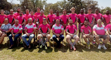 Several rugby players dressed in matching pink shirts and white pants, posing together in a relaxed environment.