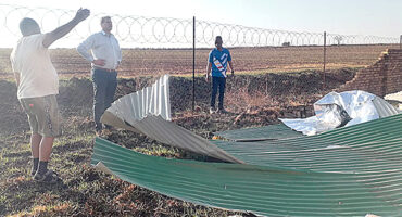 Three men stand near a fence under a metal roof, engaged in conversation outdoors.