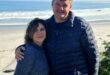A man and woman stand together on a sandy beach, with gentle ocean waves in the background.