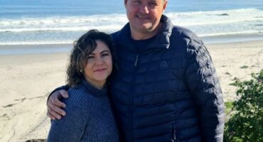A man and woman stand together on a sandy beach, with gentle ocean waves in the background.