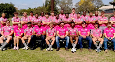 A group of men wearing matching pink shirts and shorts, standing together outdoors, smiling at the camera.