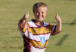A young boy stands on a field, smiling and giving a thumbs up gesture