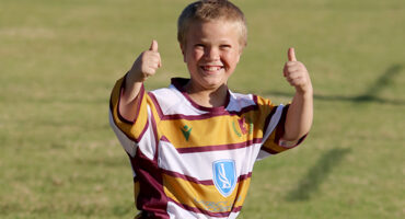 A young boy stands on a field, smiling and giving a thumbs up gesture