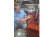 A man sits in a room surrounded by various tools, focused on a project in front of him.
