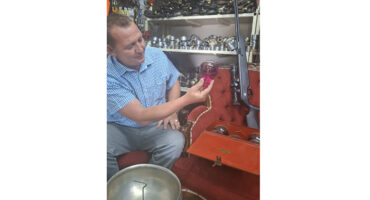 A man sits in a room surrounded by various tools, focused on a project in front of him.