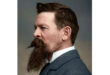 A well-dressed man with a beard wearing a suit, standing confidently with a neutral expression.