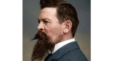 A well-dressed man with a beard wearing a suit, standing confidently with a neutral expression.