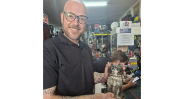 A man with glasses and a beard holds a small statue, smiling as he showcases the intricate details of the figure.