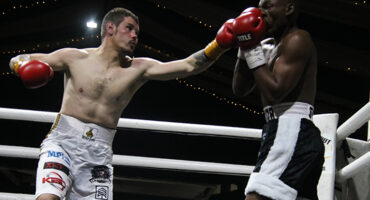 Two boxers engage in a match inside a boxing ring, illuminated by bright lights at night.