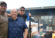 Three men stand beside a braai, engaged in conversation while preparing food outdoors.