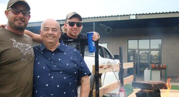 Three men stand beside a braai, engaged in conversation while preparing food outdoors.