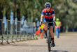 A female cyclist rides down a road, wearing a helmet and cycling gear, with trees lining the path.