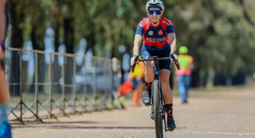 A female cyclist rides down a road, wearing a helmet and cycling gear, with trees lining the path.