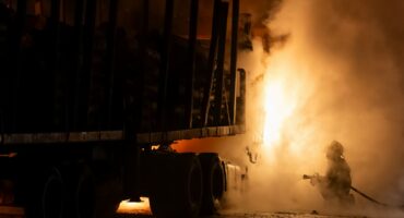 A firefighter battles a blazing truck carrying logs on a highway at night, with dense smoke and orange flames obscuring much of the scene.