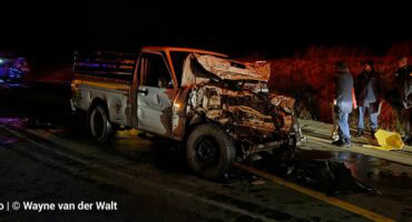 A severely damaged white bakkie with a crumpled front end sits on a dark highway at night. Several emergency personnel stand near the vehicle, with debris and liquid visible on the asphalt.