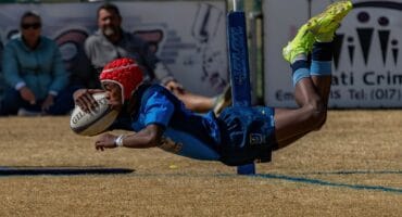 'n Rugbyspeler van Ligbron in 'n rooi helm en blou uniform duik deur die lug om 'n drie te druk.