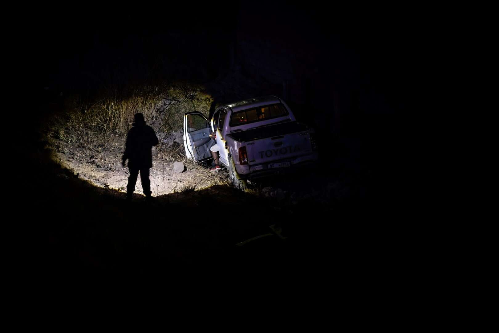 A white bakkie is seen where it came to a standstill at the bottom of a dark, grassy embankment at night. A person stands silhouetted by a strong light beam directed towards the vehicle, near the open driver's side door.