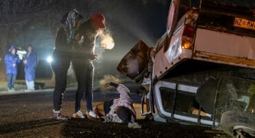 An overturned white bakkie lies on its roof on the side of a road at night. Two people stand near a third person who is lying on the ground. In the background, two more people in blue raincoats stand by. The scene is lit by artificial light.