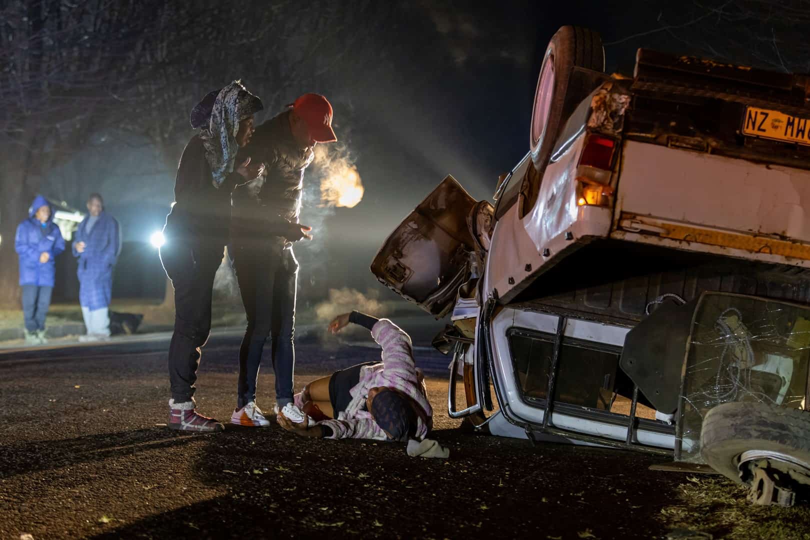 An overturned white bakkie lies on its roof on the side of a road at night. Two people stand near a third person who is lying on the ground. In the background, two more people in blue raincoats stand by. The scene is lit by artificial light.