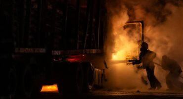 Firefighters battle a blazing truck carrying logs on a dark highway, with thick smoke filling the air.