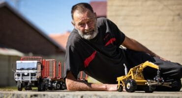 Ouer man met ’n grys baard lê op die grond tussen twee gedetailleerde handgemaakte miniatuurmodelle van ’n vragmotor en ’n Bell-bosbouvoertuig.