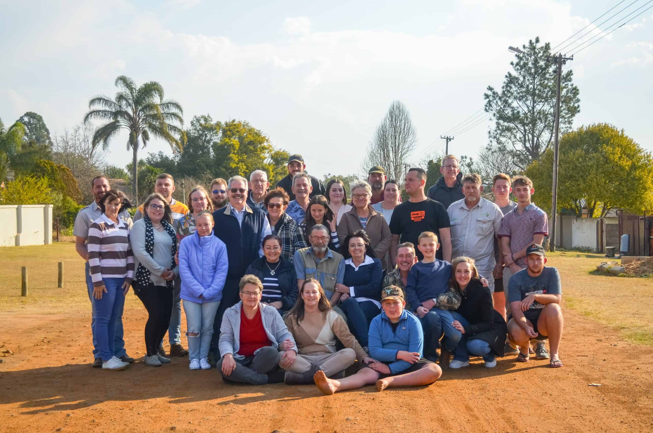 'n Groot groep mense, van verskillende ouderdomme, pos vir 'n familiefoto buite op 'n oop, grond-terrein.
