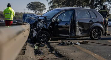 Black hatchback car with severe front and side damage after collision with guardrail on a highway, debris on road, and two emergency responders in the background.