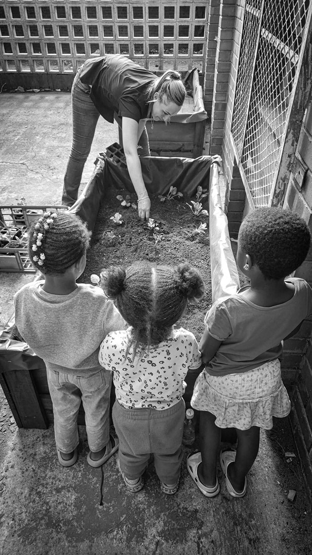 Aspirant Knight Theressa van der Walt plants vegetable seedlings while young children from Children of the Dawn watch with curiosity.