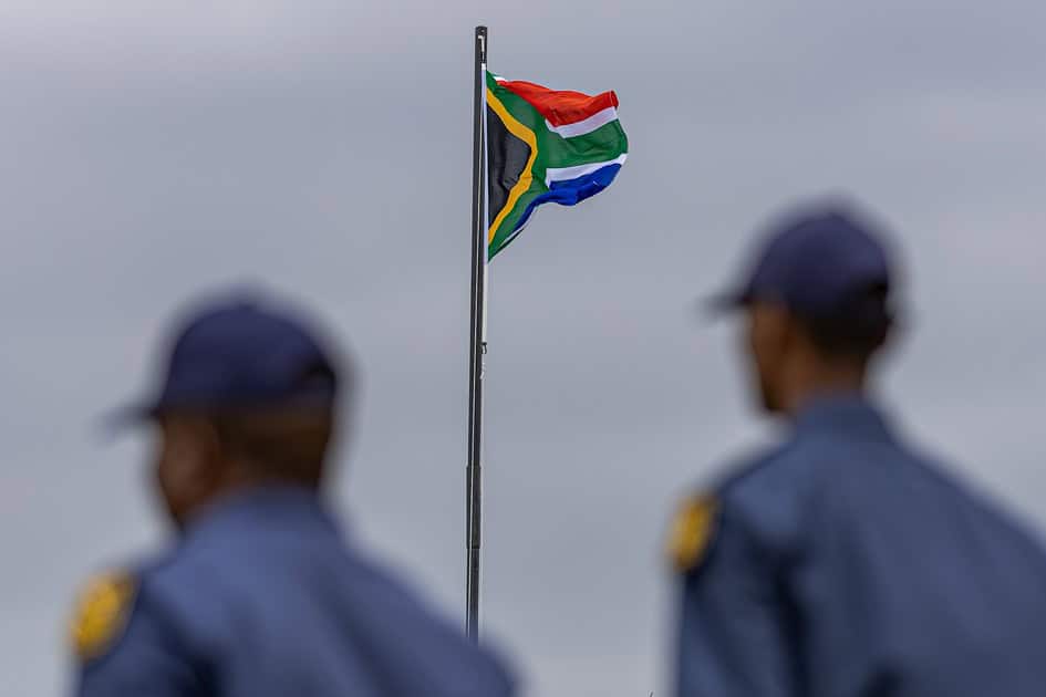 The South African flag flutters during the Safer Festive Season parade in Ermelo, framed between two blurred police officers in the foreground.