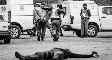 Police officer lies on the ground after being shot during a simulated robbery, while another officer arrests the suspect and escorts him to a police van in Ermelo.