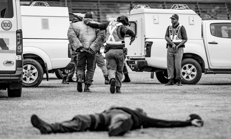 Police officer lies on the ground after being shot during a simulated robbery, while another officer arrests the suspect and escorts him to a police van in Ermelo.