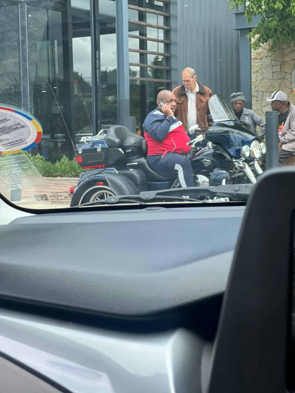 Former MEC Vusi Shongwe sitting on a stationary motorcycle at Merino Mall, talking on his phone while wearing a tracksuit.