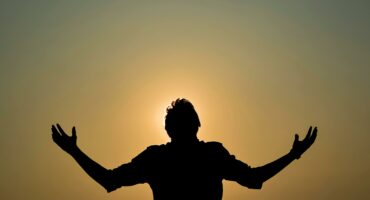 Silhoeët van ’n man wat sy hande in die lug lig teen ’n ligte agtergrond.