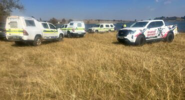Emergency response vehicles, including SAPS police and a Spartan bakkie, parked on a grassy field next to Douglas Dam in Ermelo during a reported drowning response.