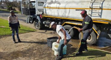 Inwoners vorm rye langs Generaal Bothastraat terwyl water uit ’n konstruksievoertuig uitgedeel word te midde van voortdurende wateronderbrekings in Ermelo.