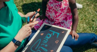 Adult assisting a young child with learning activities outdoors using a chalkboard