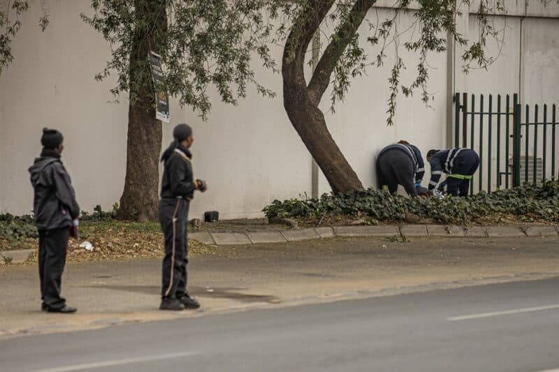 Forensic investigators examine the body of an unidentified man found on a sidewalk behind vegetation near a palisade fence at Oak Centre in Ermelo.