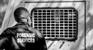 Forensic services officer stands near a detained man inside a police vehicle.