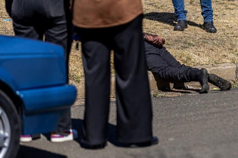 Detained man lies beside the kerb near a blue vehicle as people stand nearby.