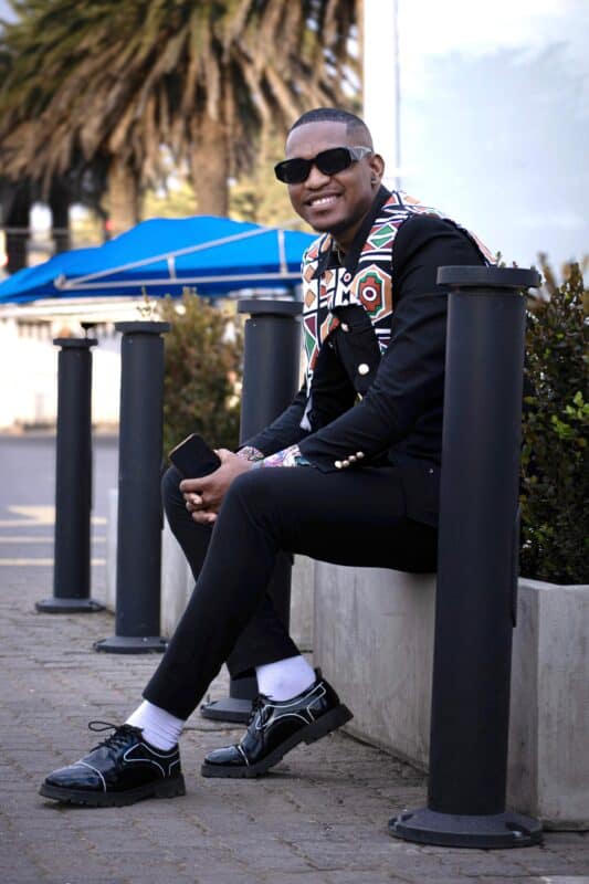 Ermelo presenter Siphamandla Nkosi seated outdoors in a black outfit with colourful patterned detailing.
