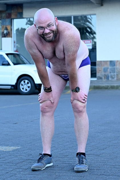 A smiling Daredevil Run participant in purple speedos leans forward with his hands on his knees.