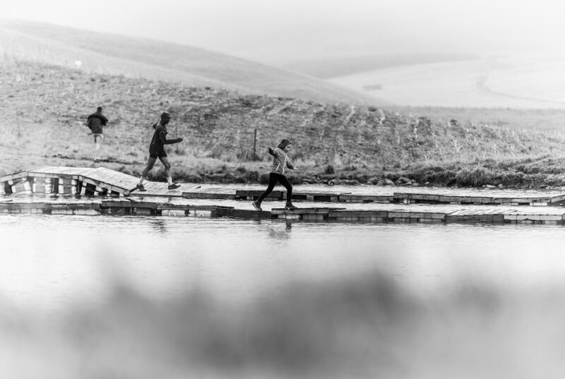 Drie deelnemers steek die waterbrug oor tydens die Glen Aggy Trail Run in 2025.