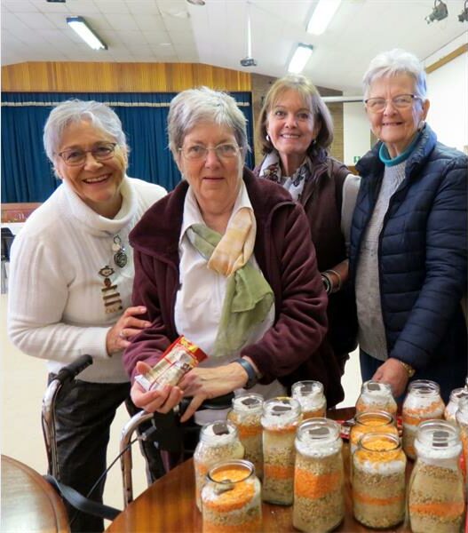Four women are standing around a table with jars of food.