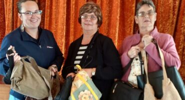 Three women are holding bags and smiling.