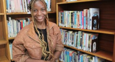 A woman with dreadlocks sitting in front of a bookshelf.
