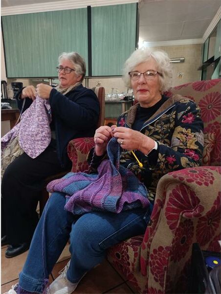 Two older women sitting in chairs knitting.
