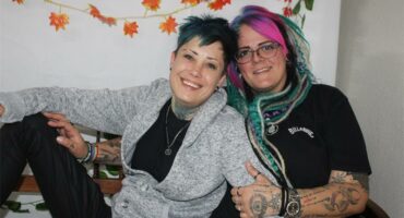 Two women with colorful hair and tattoos sitting on a bench.