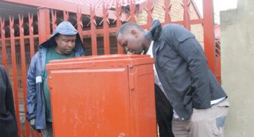 Two men looking at an orange electric box in front of a fence.